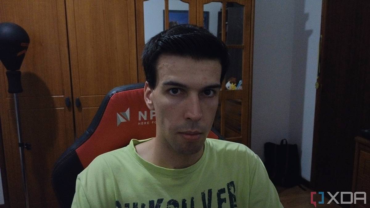A picture of a man sitting on a chair with furniture behind him. The picture was taken with the Insta360 Link webcam with the HDR feature enabled, and it looks much darker overall