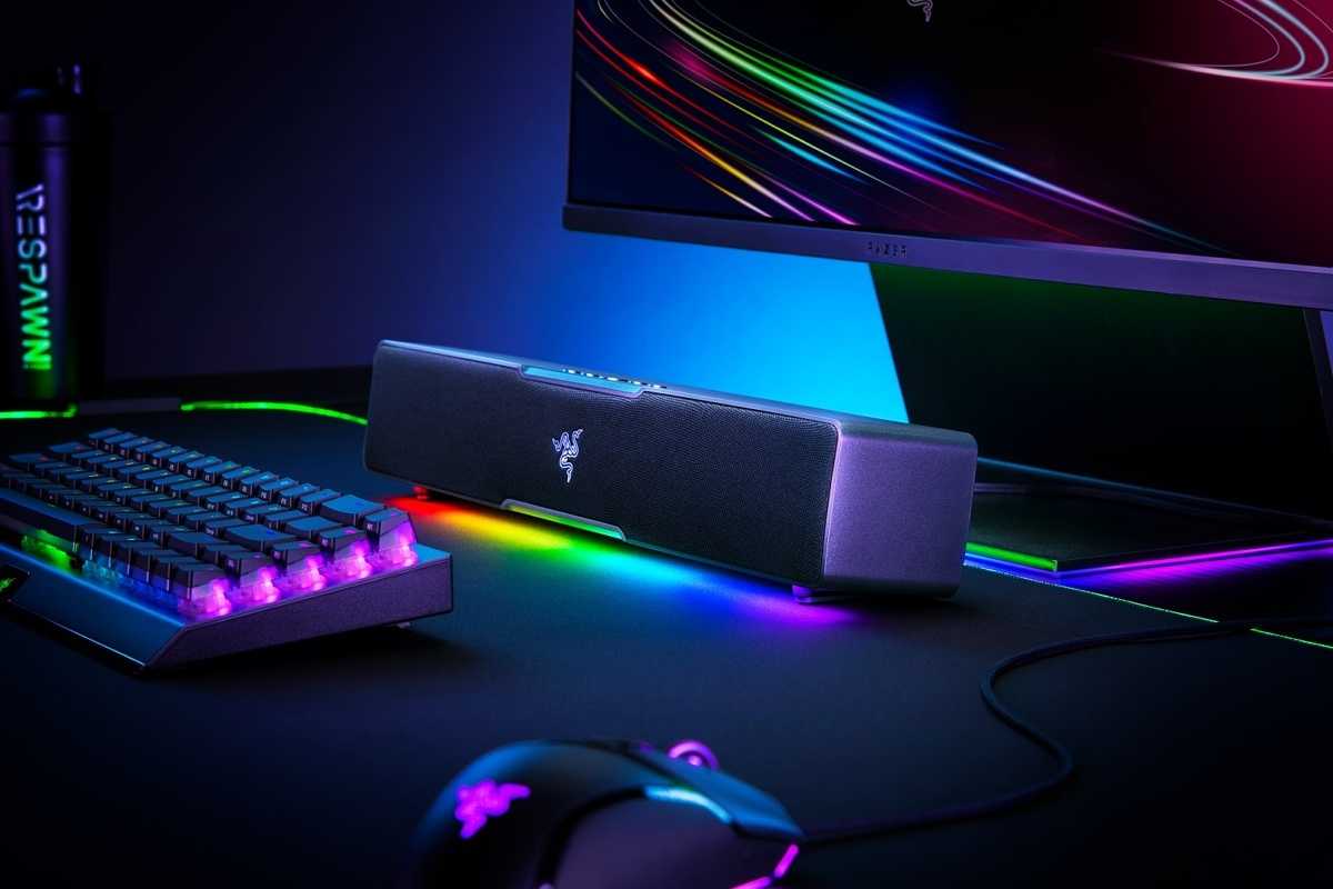 Angled view of the Razer Leviathan V2 X soundbar with RGB lighting enabled, next to other peripherals including a keyboard, mouse, and monitor.