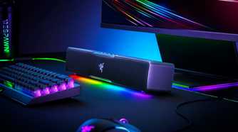 Angled view of the Razer Leviathan V2 X soundbar with RGB lighting enabled, next to other peripherals including a keyboard, mouse, and monitor.