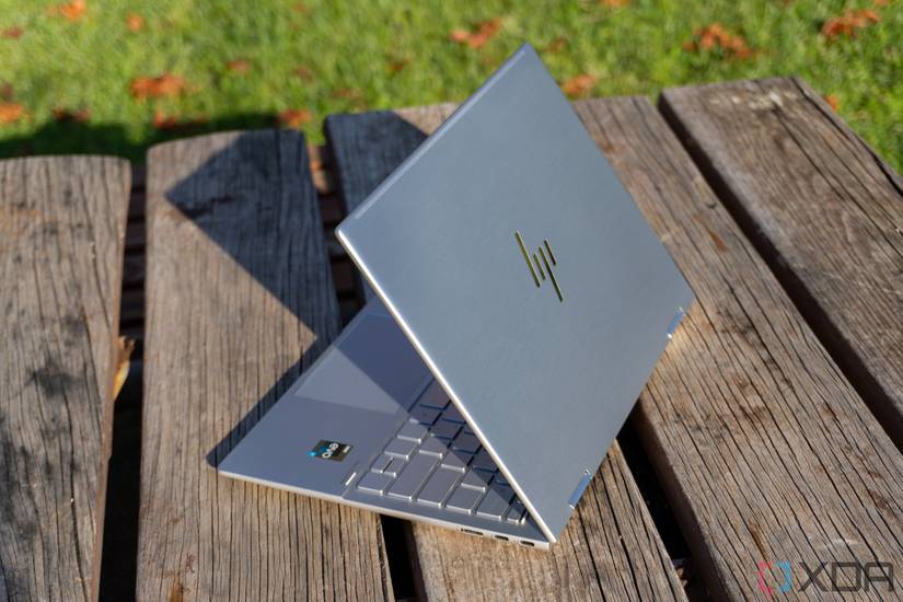 The HP Envy x360 13 seen at a right-side angle with the lid open at about 60 degrees. The laptop is sitting on a wooden table with grass in the background