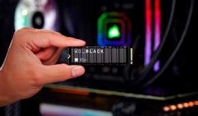 A person holding the WD Black SN850X gaming ssd with their thumb and the index finger with a gaming PC with RGB lights in the background.