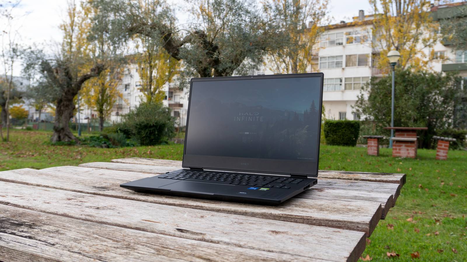The Omen 16 laptop sitting on a wooden board table in an outdoor garden. The laptop is displaying the title screen for Halo Infinite, and various vegetation can be seen around the table where the laptop is