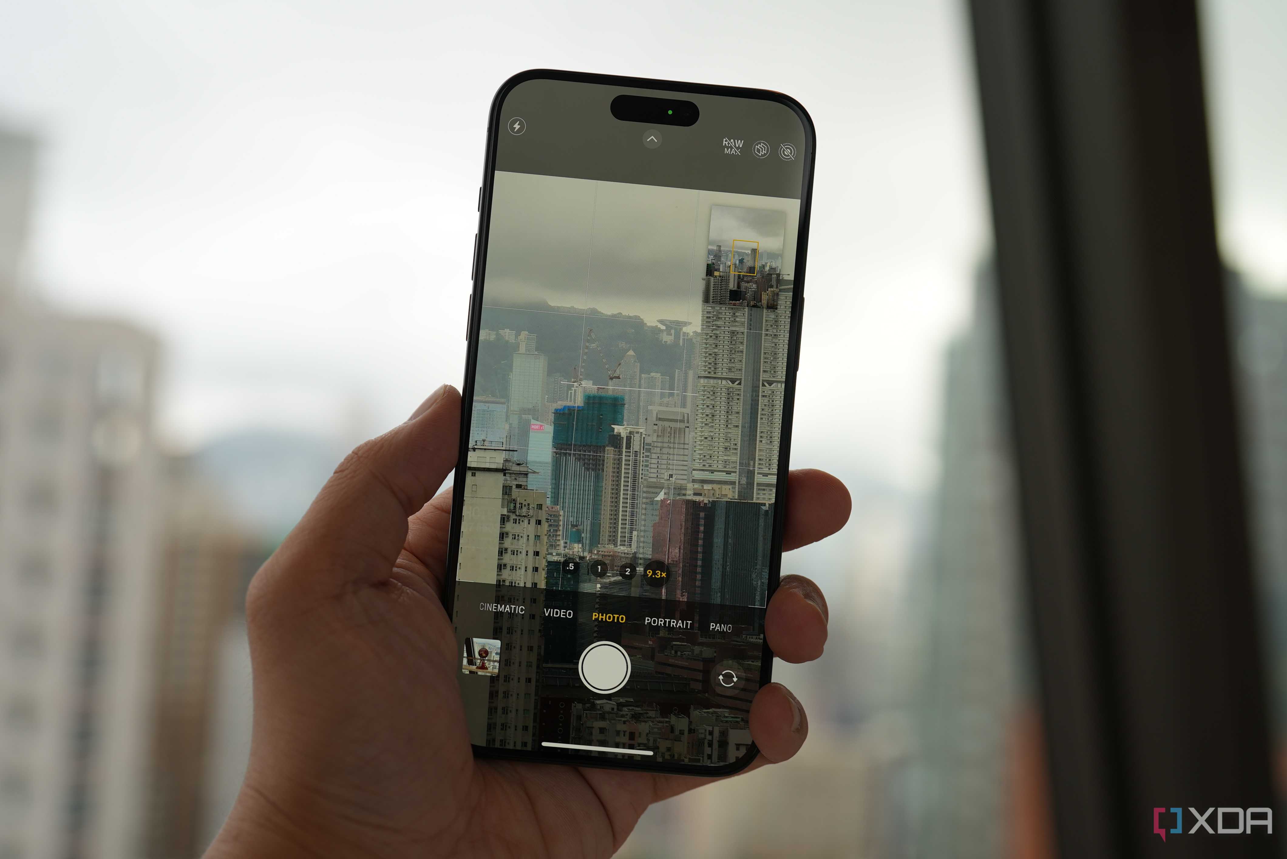 An iPhone 15 Pro being held in a hand taking a photo of a dense city full of high-rises on a cloudy day through a window.