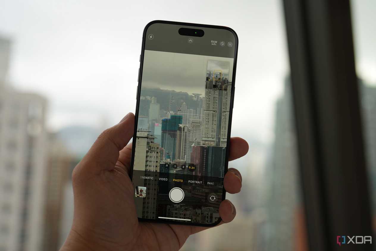 An iPhone 15 Pro being held in a hand taking a photo of a dense city full of high-rises on a cloudy day through a window.