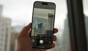 An iPhone 15 Pro being held in a hand taking a photo of a dense city full of high-rises on a cloudy day through a window.