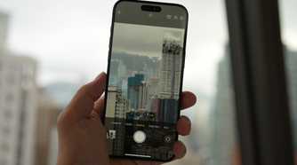 An iPhone 15 Pro being held in a hand taking a photo of a dense city full of high-rises on a cloudy day through a window.