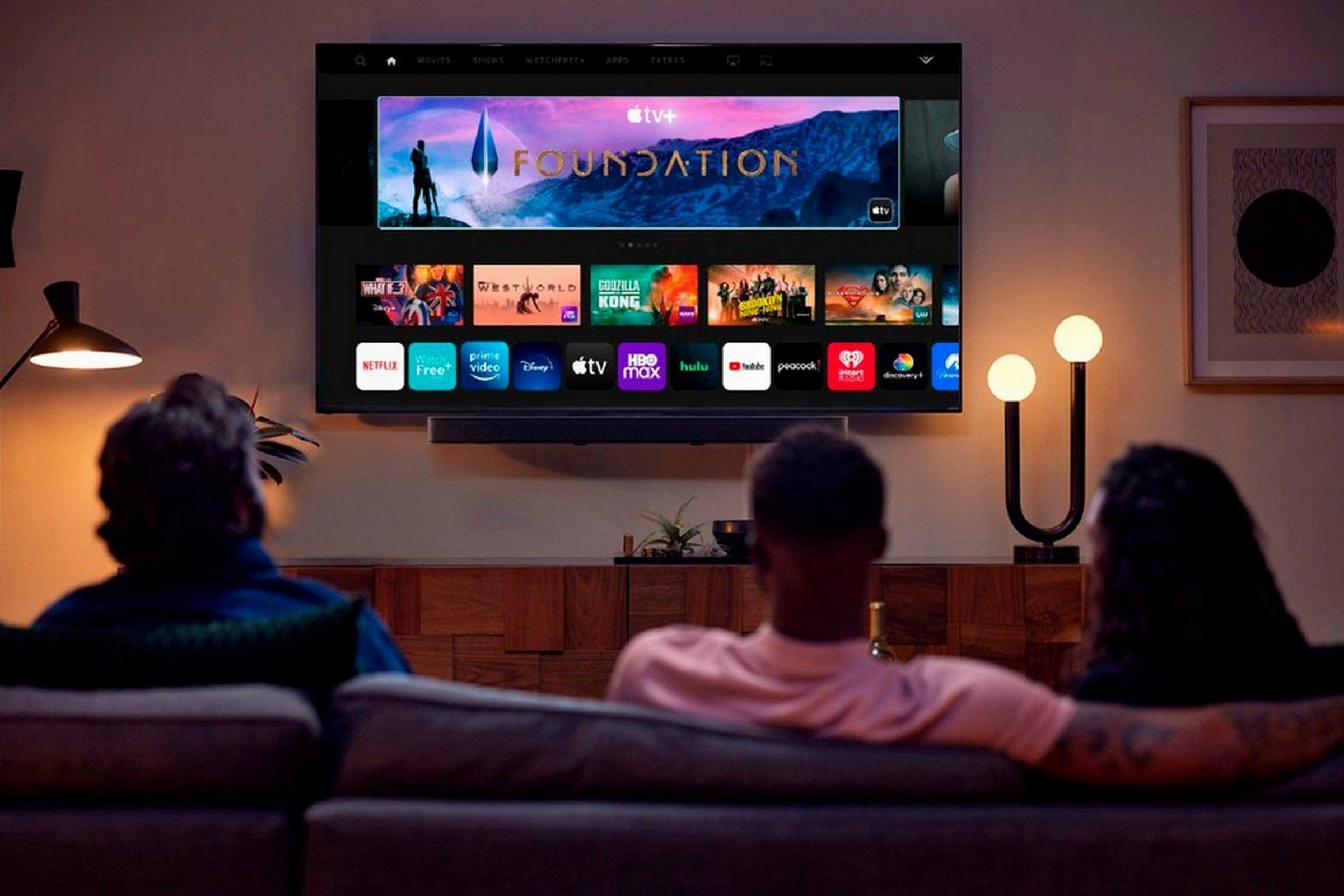 Three people on a couch, pictured from the back, watching a TV mounted on the wall