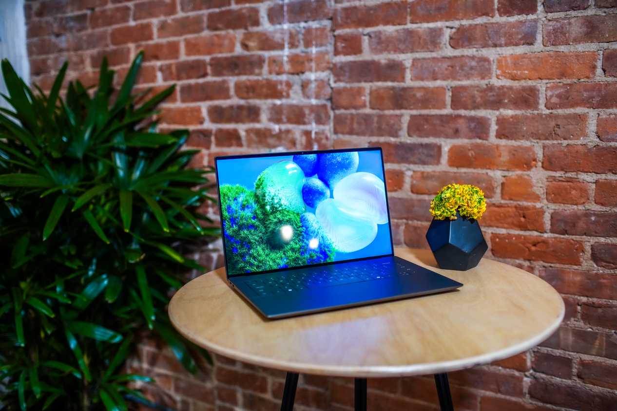 A Dell XPS 14 laptop sitting on a table next to a vase filled with flowers in front of a brick wall with a green plant in the background.