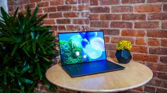 A Dell XPS 14 laptop sitting on a table next to a vase filled with flowers in front of a brick wall with a green plant in the background.