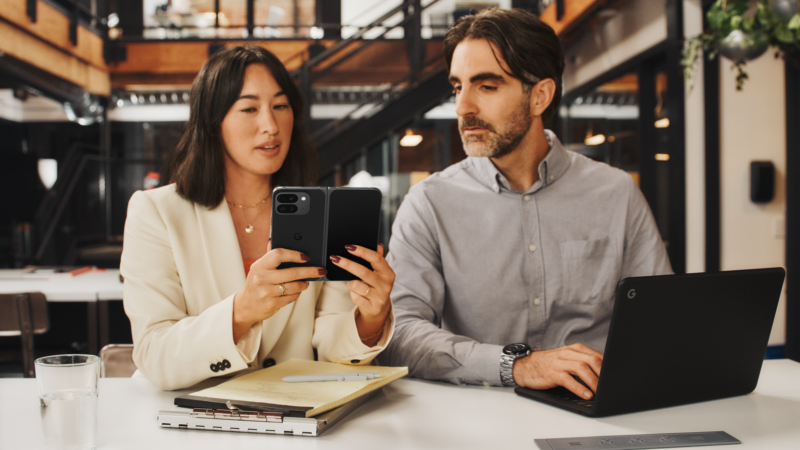 A woman holding a Google Pixel 9 Pro Fold and showing it to a man next to her, who has a laptop in front of him