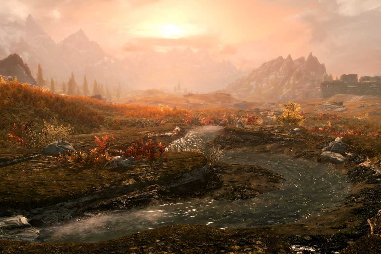A screenshot from the game The Elder Scrolls V: Skyrim, showing a beautiful sunset as small stream of water runs through a lush field bathed in the light of the setting sun.