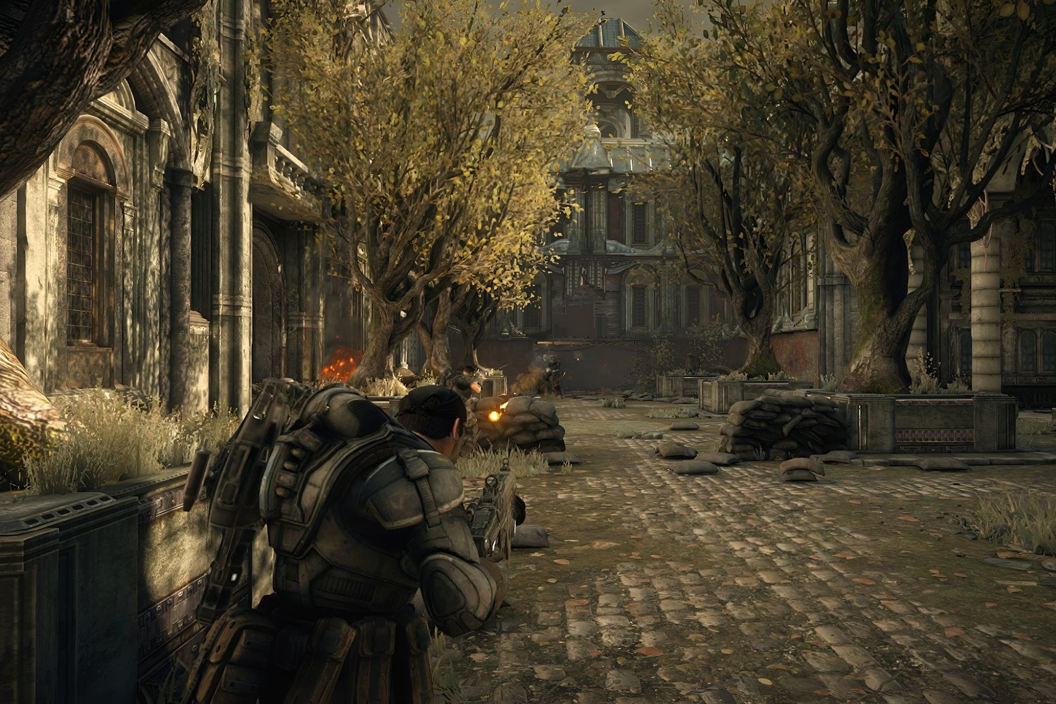 A screenshot from the game Gears of War: Ultimate Edition, showing a man in bulky armor standing in a paved garden with trees on either side.