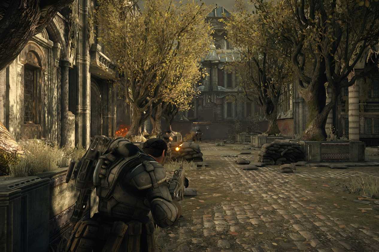A screenshot from the game Gears of War: Ultimate Edition, showing a man in bulky armor standing in a paved garden with trees on either side.