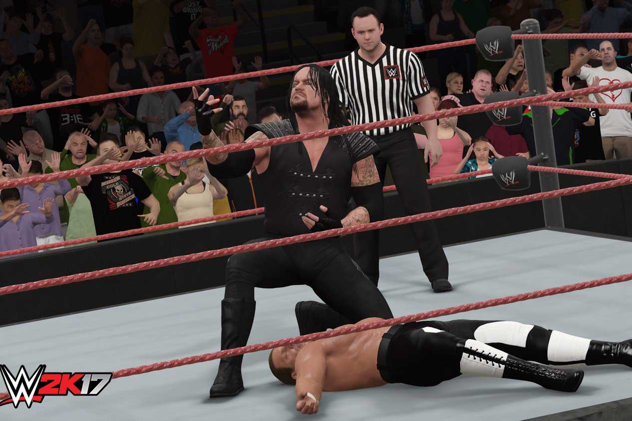 A screenshot from WWE 2K17, showing the Undertaker, in black overalls, lean over a knocked out Hunter Hearst Helmsley.