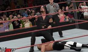 A screenshot from WWE 2K17, showing the Undertaker, in black overalls, lean over a knocked out Hunter Hearst Helmsley.
