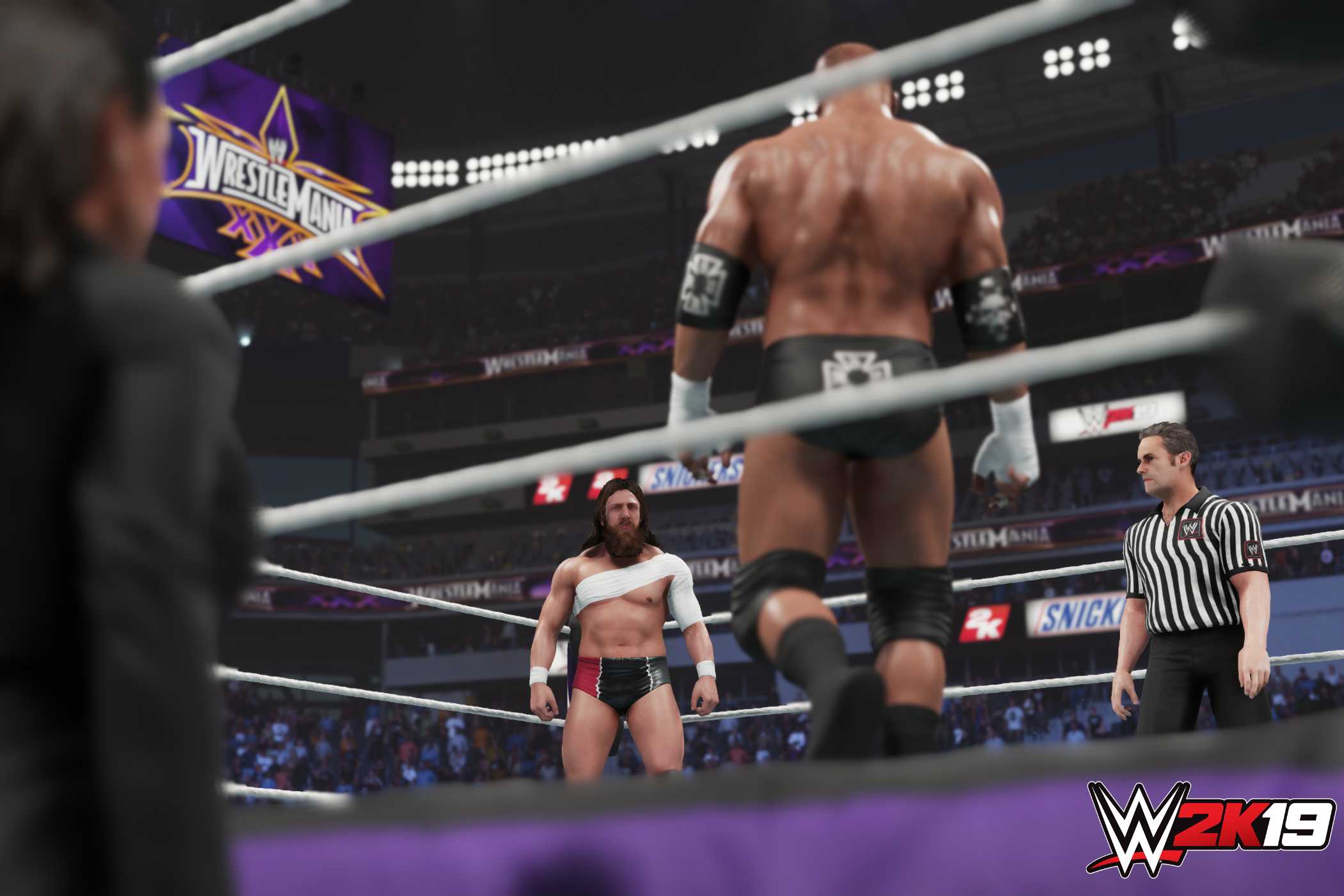 A screenshot from WWE 2K19, showing a bald Triple H face off against a bandaged Daniel Bryan in the ring as a referee looks on inside the ring.