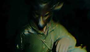 An image from the game Alan Wake II, showing a person in a green overcoat and brown leather gloves wearing a deer mask.