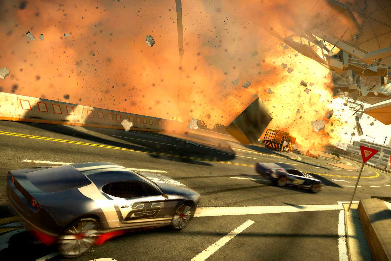An image from the game "Split Second", showing cars race alongside a stadium while it blows apart and goes up in flames.