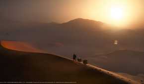 An image from the game Death Stranding 2: On The Beach, showing Sam walking along a sand dune as the sun sets.