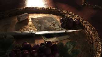 An image from the game Mafia: The Old Country, showing a bloodied knife sitting on a gold plate next to some grapes and a blood-soaked photograph.