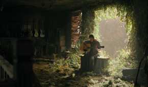 A screengrab from episode 4 of the HBO show The Last of Us Season 2, showing Ellie playing a guitar while seated in sunlight.