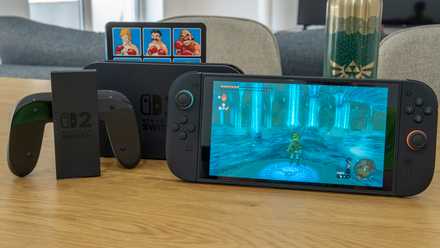A Nintendo Switch 2 console, its dock, and the Joy-Con 2 grip, all standing next to each other on a wooden table. The console is running The Legend of Zelda tears of the Kingdom