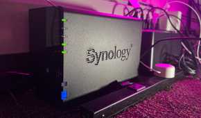A Synology NAS next to a power strip and UPS on a carpeted floor