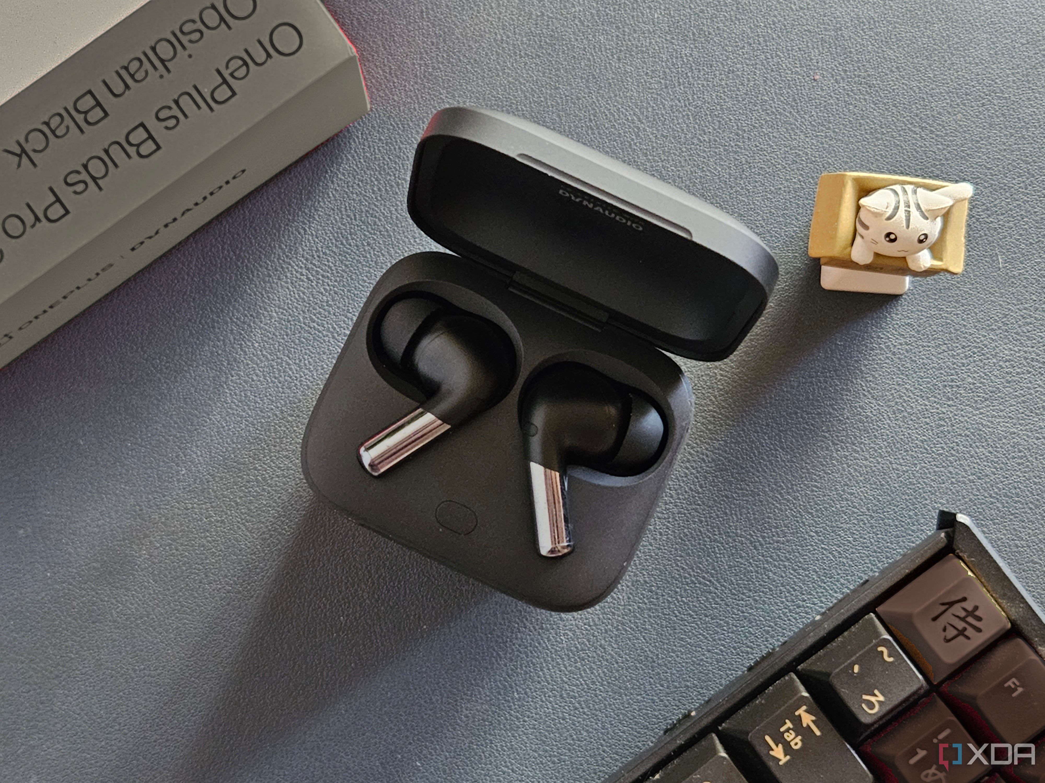 An image showing a pair of OnePlus Buds Pro 2 earbuds kept in the charging box next to its retail box, a keyboard, and an artisan keycap.