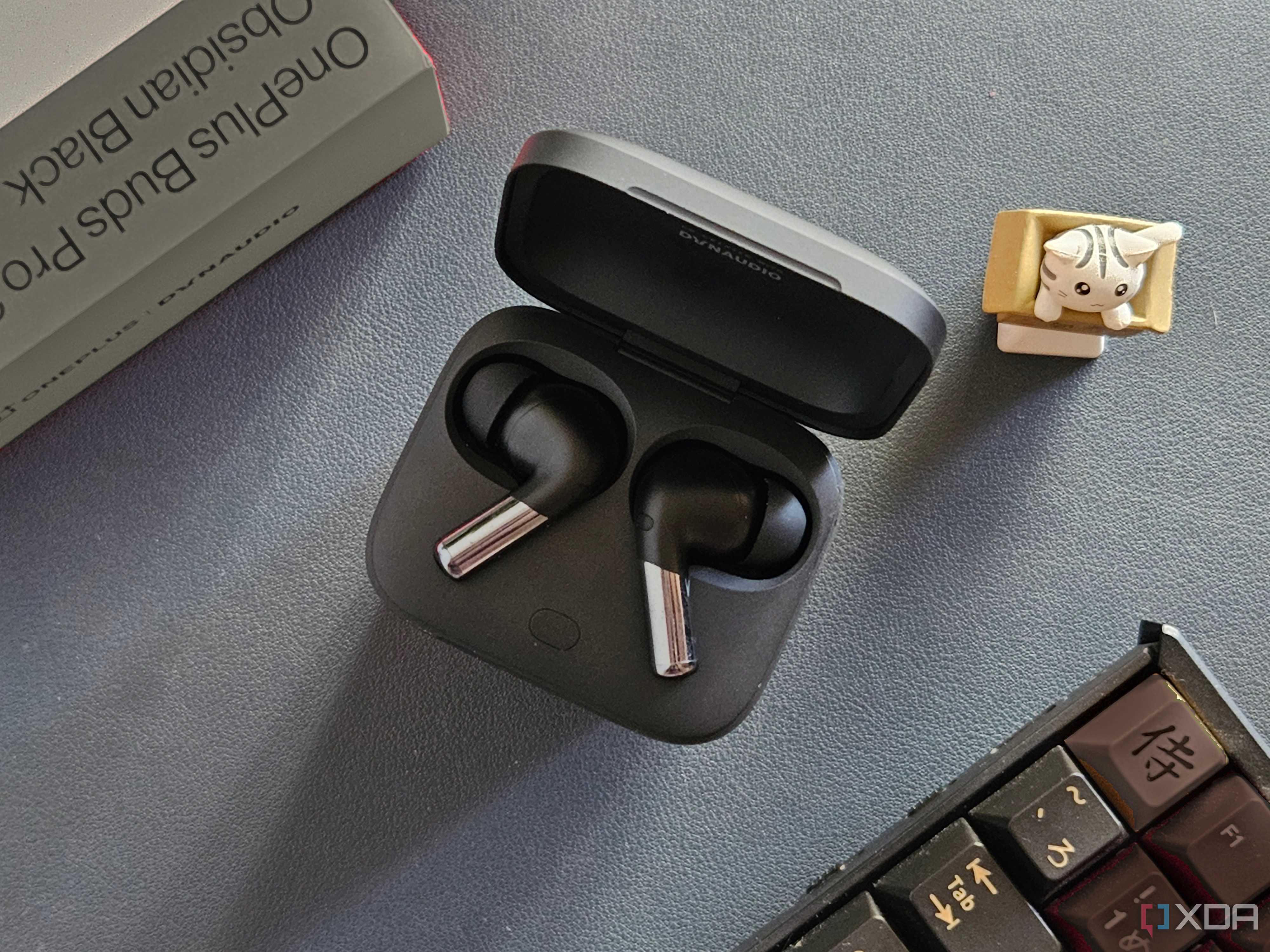 An image showing a pair of OnePlus Buds Pro 2 earbuds kept in the charging box next to its retail box, a keyboard, and an artisan keycap.