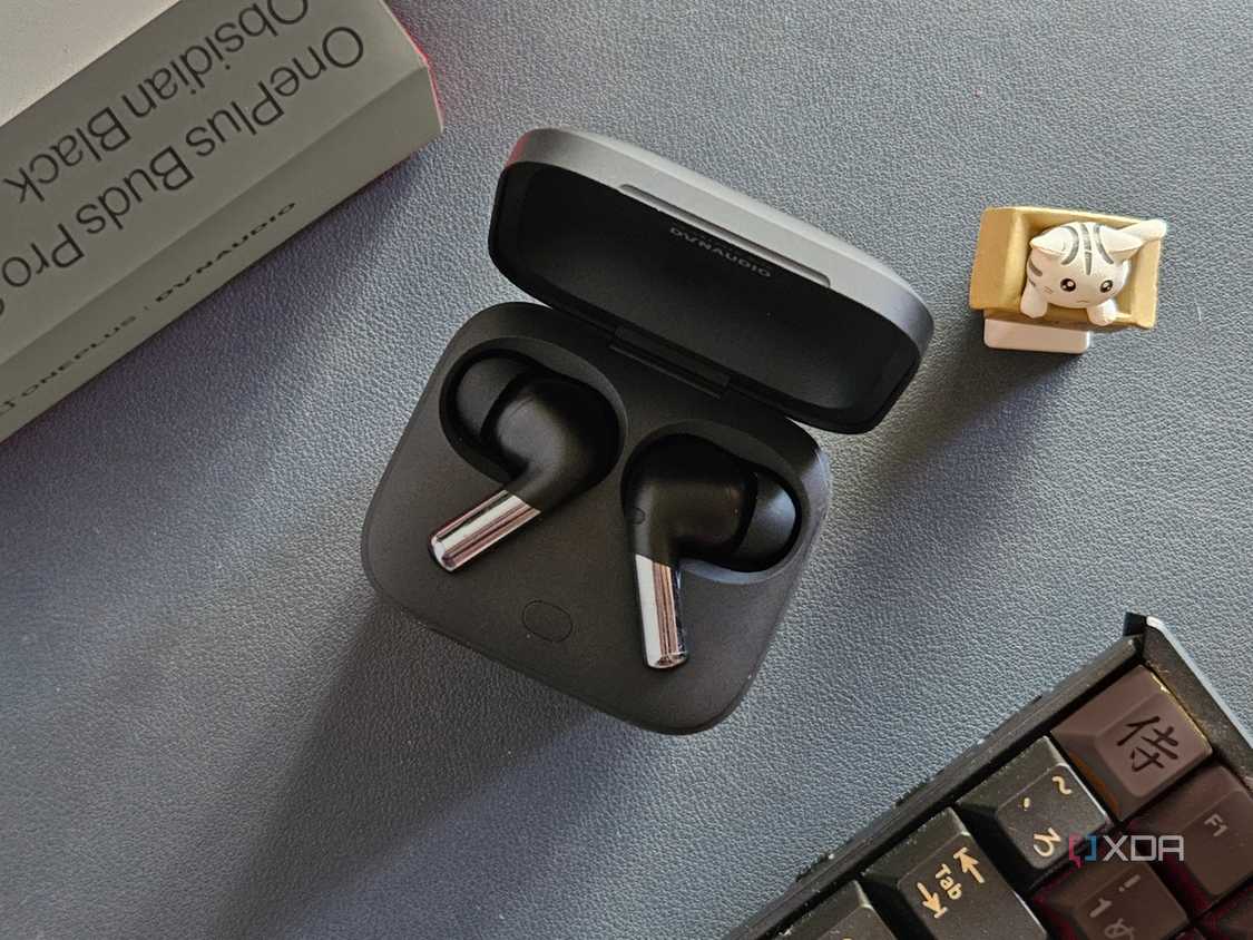 An image showing a pair of OnePlus Buds Pro 2 earbuds kept in the charging box next to its retail box, a keyboard, and an artisan keycap.