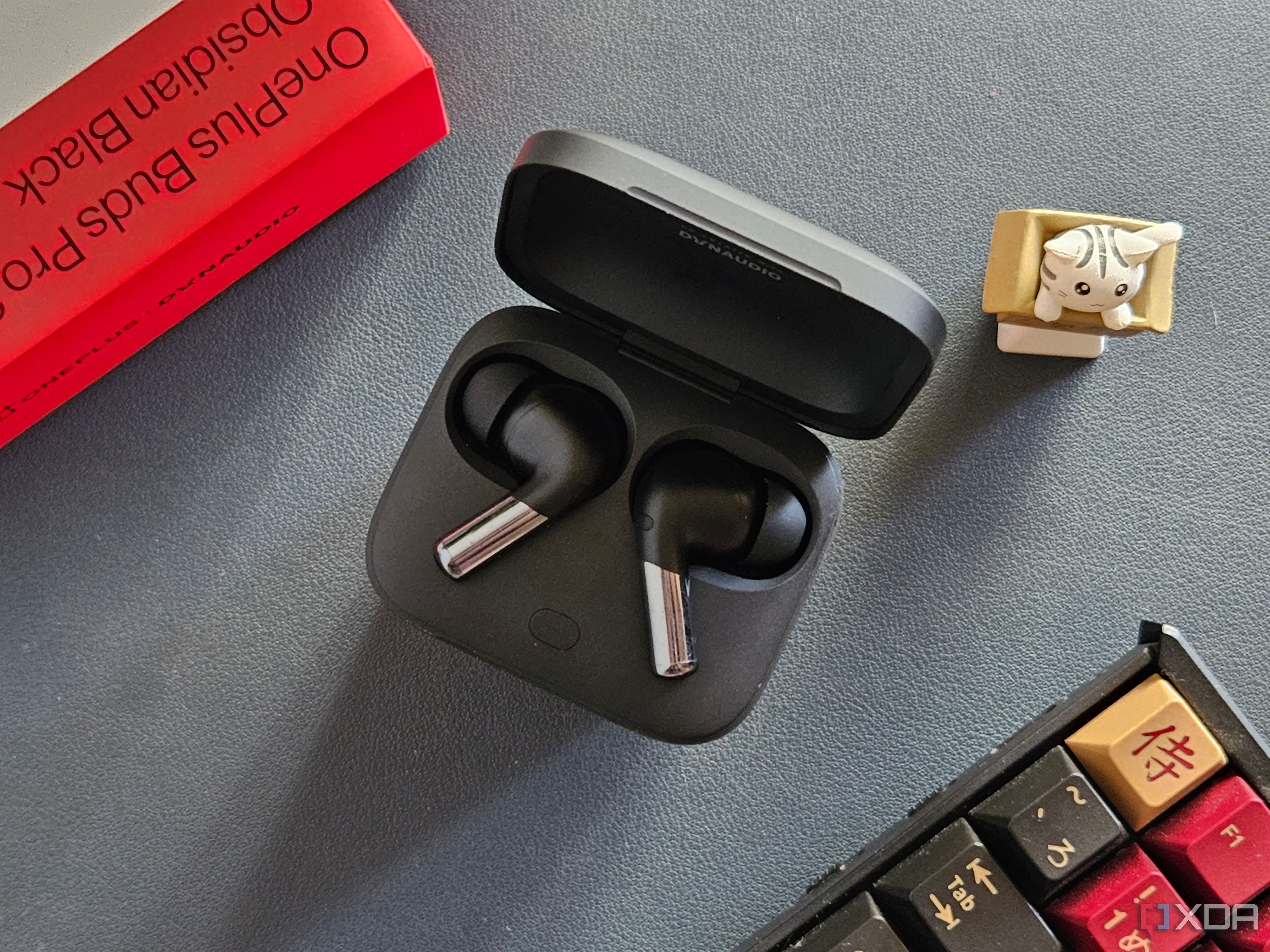 An image showing a pair of OnePlus Buds Pro 2 earbuds kept in the charging box next to its retail box, a keyboard, and an artisan keycap.