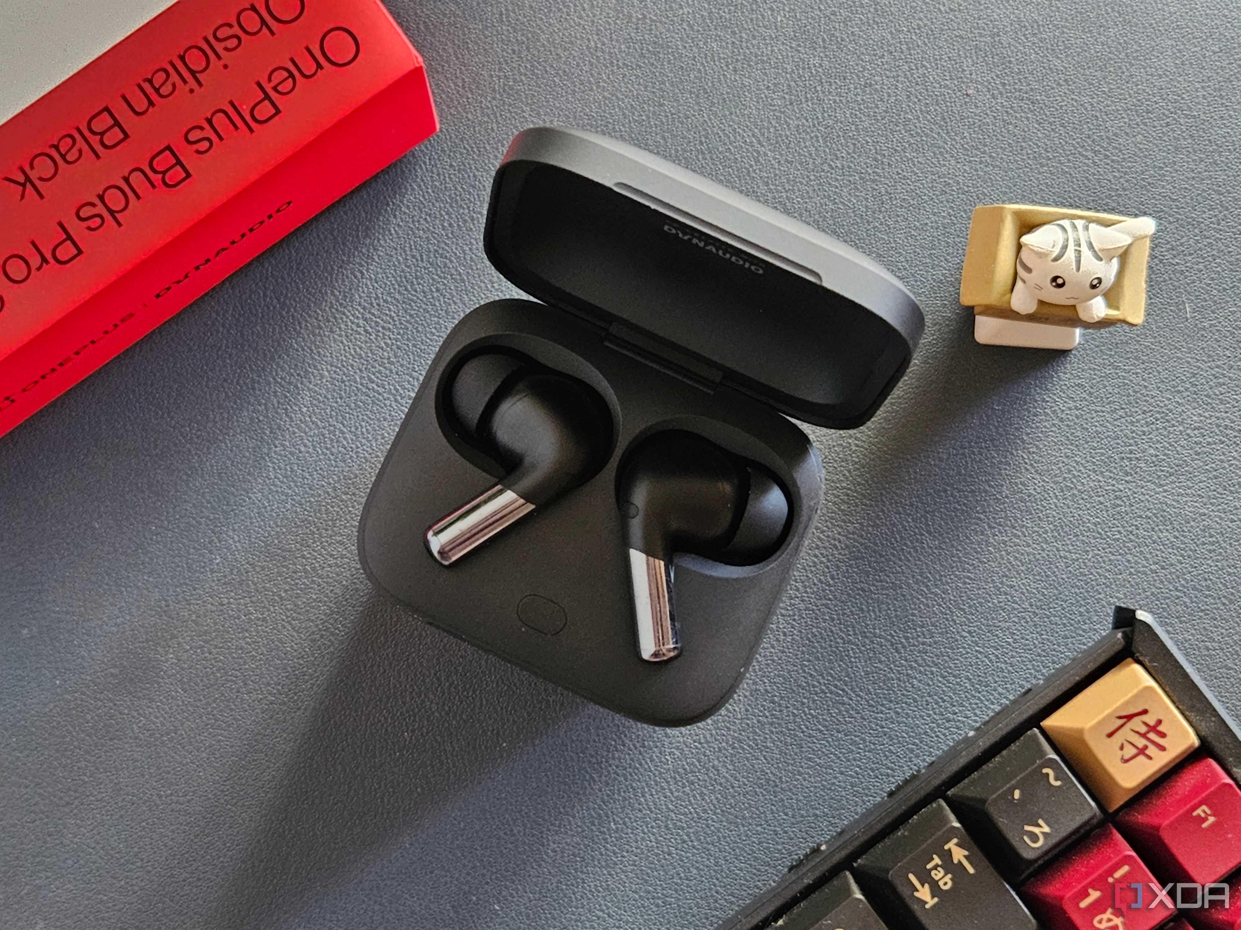 An image showing a pair of OnePlus Buds Pro 2 earbuds kept in the charging box next to its retail box, a keyboard, and an artisan keycap.