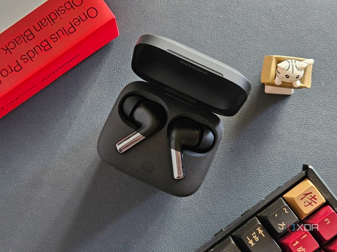 An image showing a pair of OnePlus Buds Pro 2 earbuds kept in the charging box next to its retail box, a keyboard, and an artisan keycap.