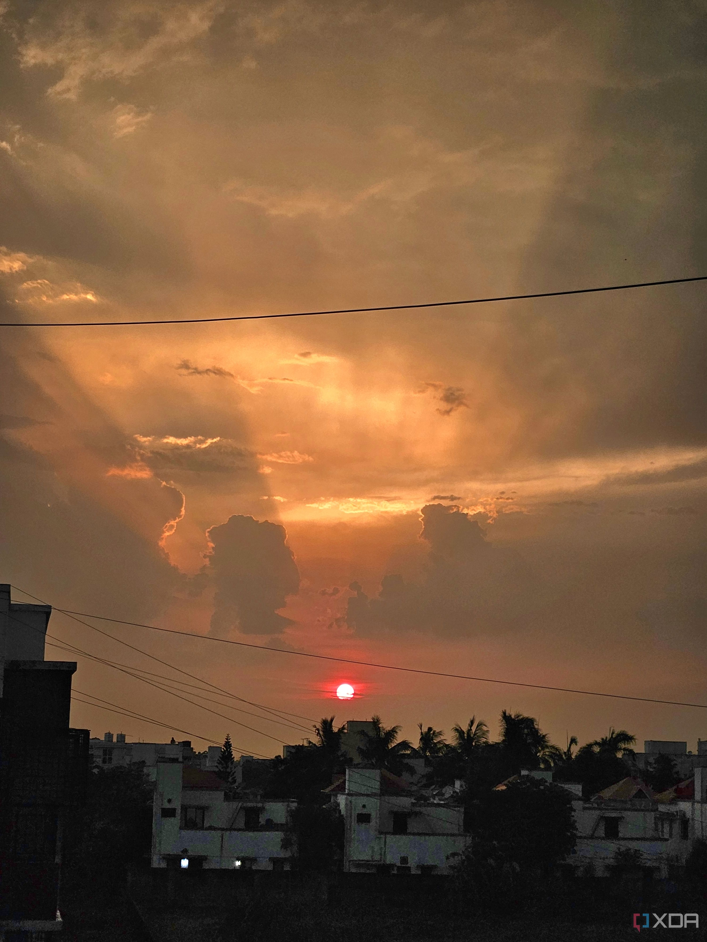 An image showing a sunset with its light shinning through the clouds, and some buildings in the foreground.