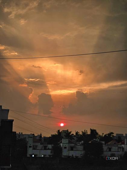 An image showing a sunset with its light shinning through the clouds, and some buildings in the foreground.