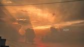 An image showing a sunset with its light shinning through the clouds, and some buildings in the foreground.