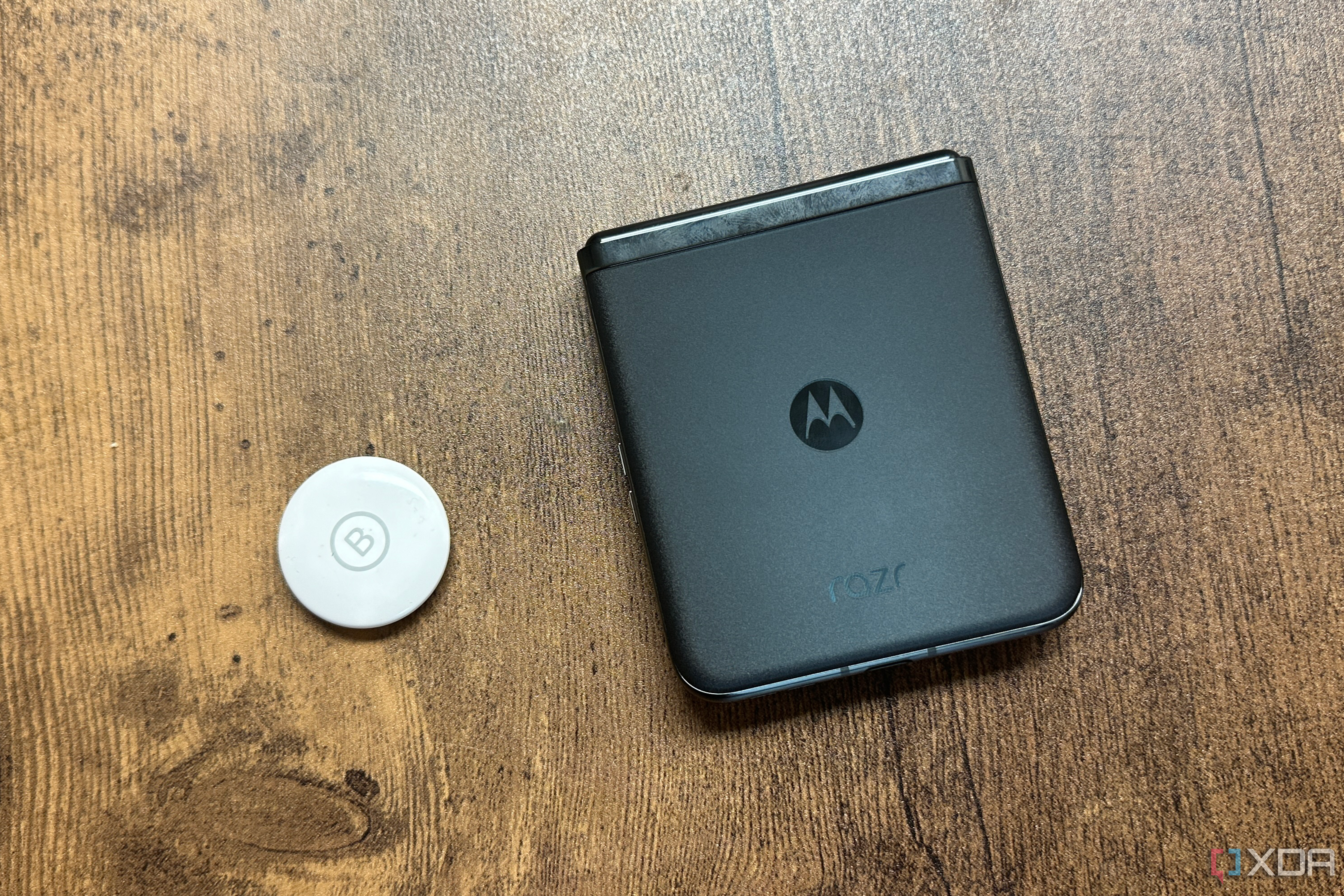 An Apple AirTag next to a Motorola Razr+ against a wood background.