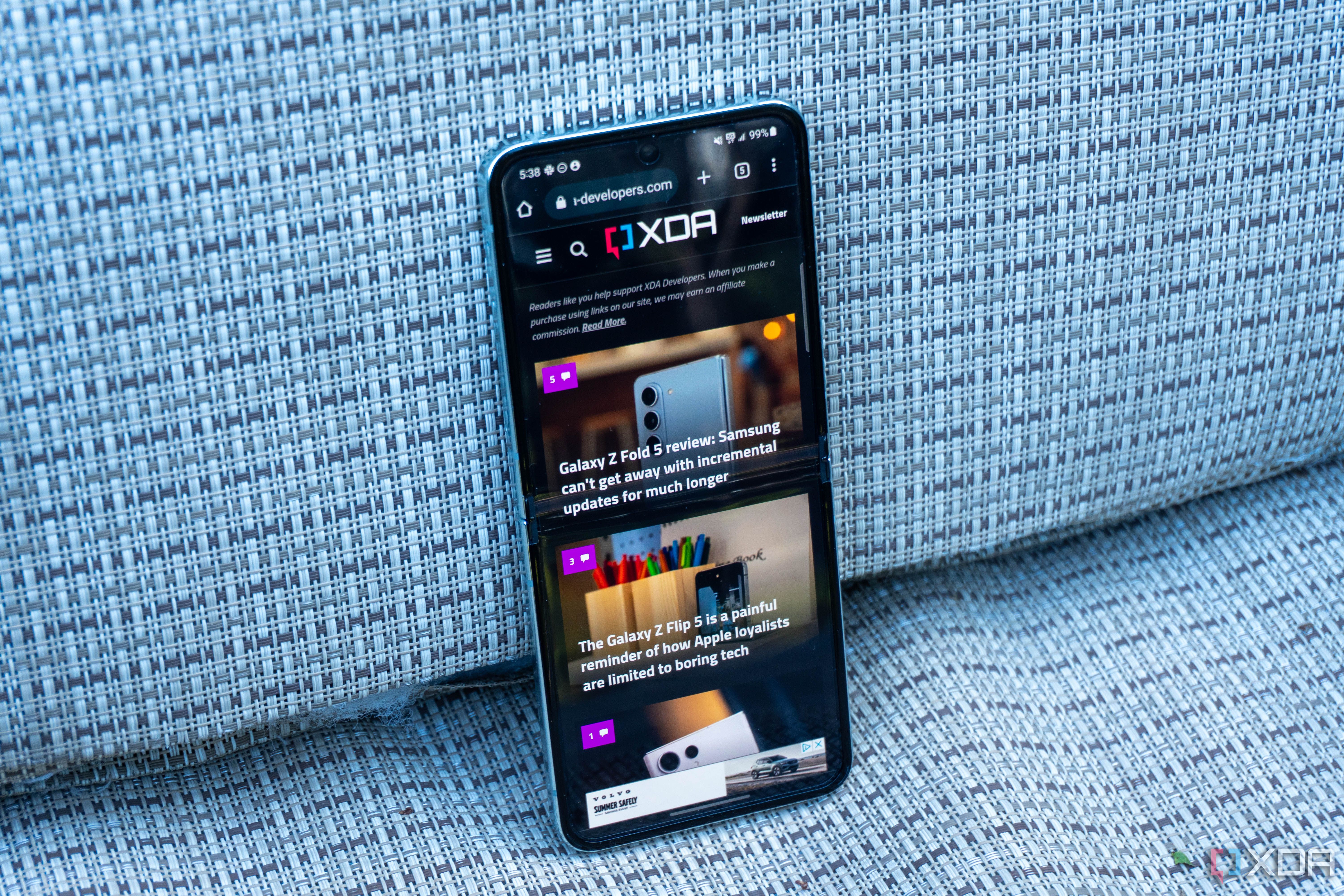A Samsung Galaxy Z Flip 5 seen from the front showing the XDA mobile website. The phone sits on a blue fabric chair outdoors.