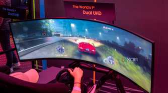 A Samsung Odyssey Neo G9 57-inch monitor displaying a racing game being played with a steering wheel.
