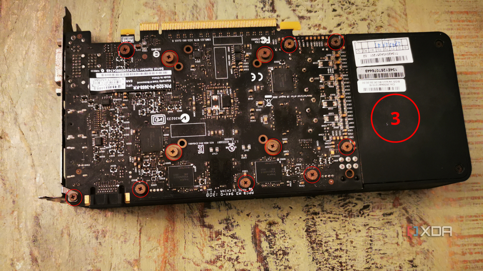 A graphics card removed from the computer, displaying the back side with a circle around each screw on the board and a numbered label for the third step.