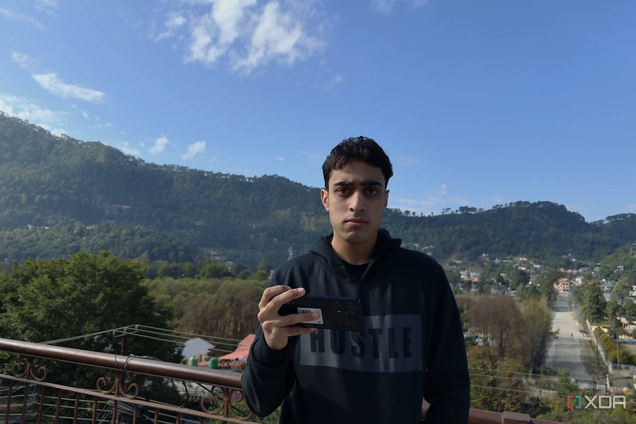A man holding a Poco F5, with a panoramic sight behind him