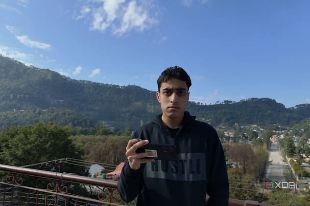 A man holding a Poco F5, with a panoramic sight behind him