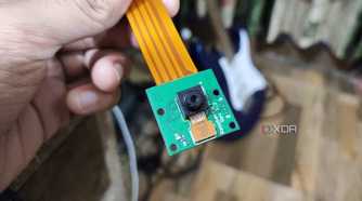 A person holding a Raspberry Pi Camera rev 1.3, with a guitar in the background