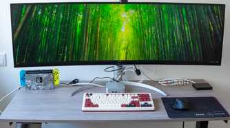 A desk setup with multiple devices, including a Geekom A8 PC under a large ultrawide monitor
