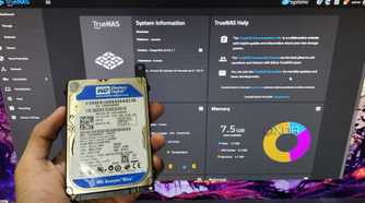 A person holding an HDD in front of a monitor displaying TrueNAS Scale web UI