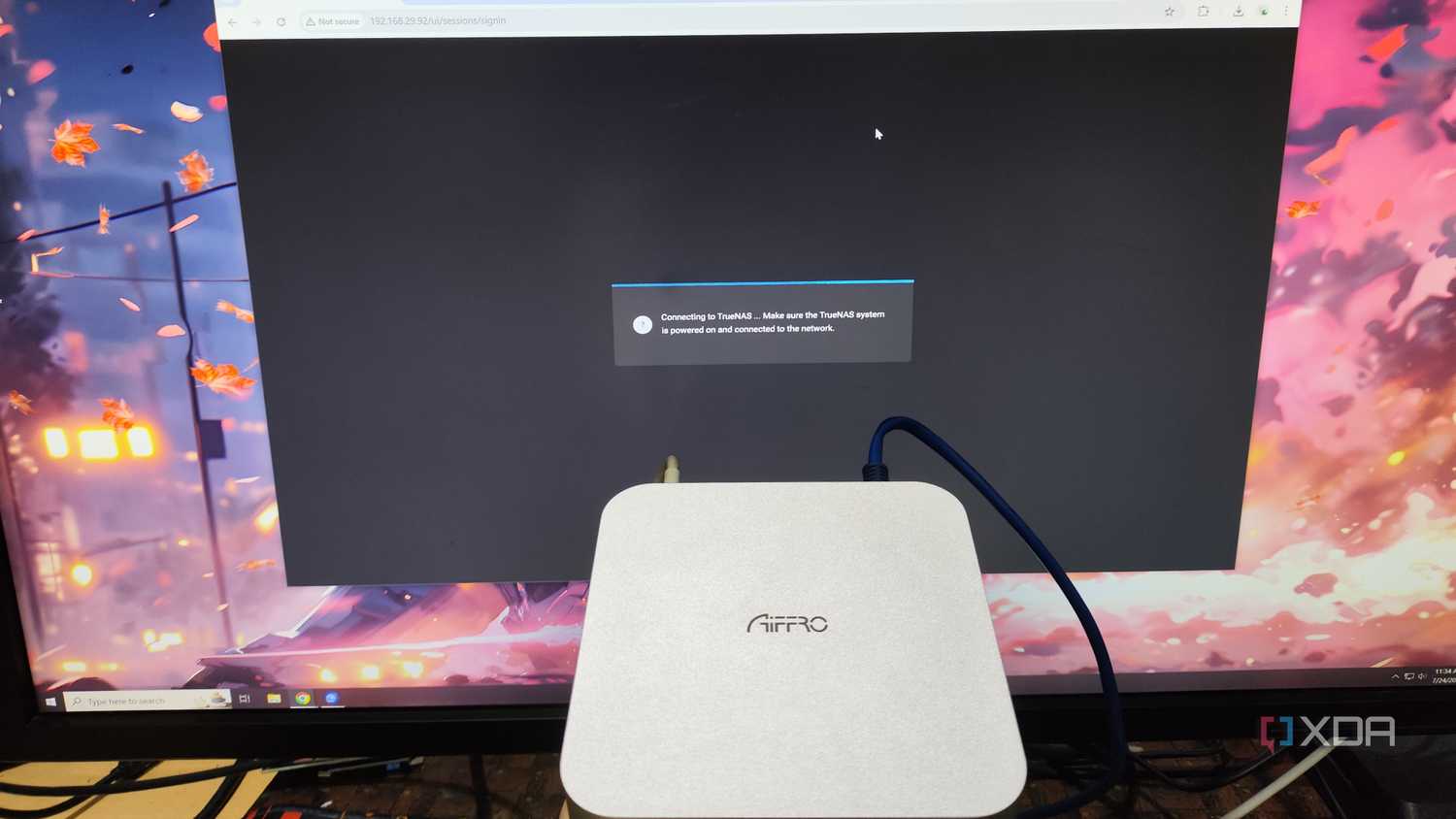 An Aiffro K100 NAS held in front of a monitor displaying the TrueNAS web UI