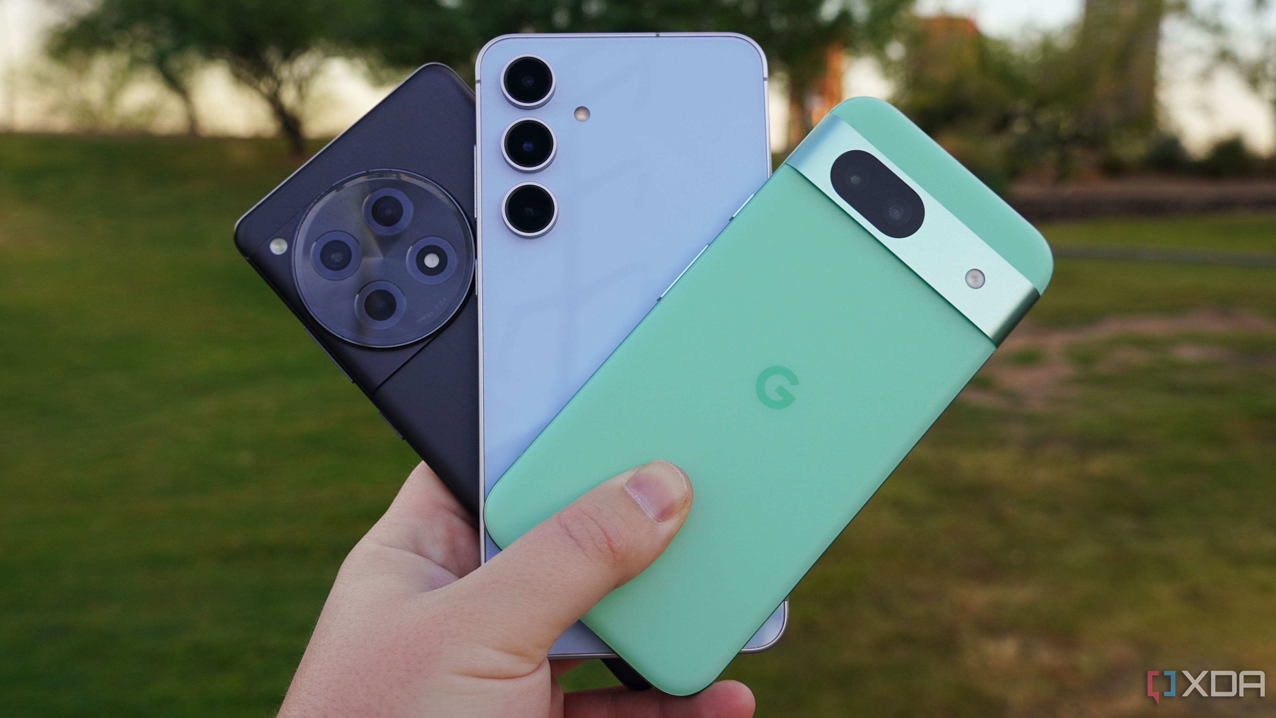 The Galaxy S24 FE in blue, the OnePlus 12R in black, and the Pixel 8a in green held in a hand against a background with green grass