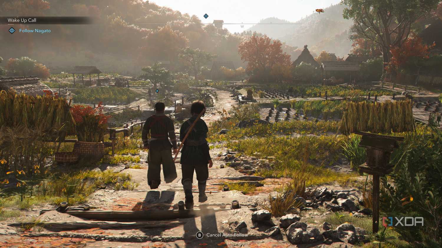 A screenshot from Assassin's Creed Shadows, showing two people walking alongside each other through a densely vegetated village.