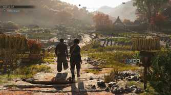 A screenshot from Assassin's Creed Shadows, showing two people walking alongside each other through a densely vegetated village.
