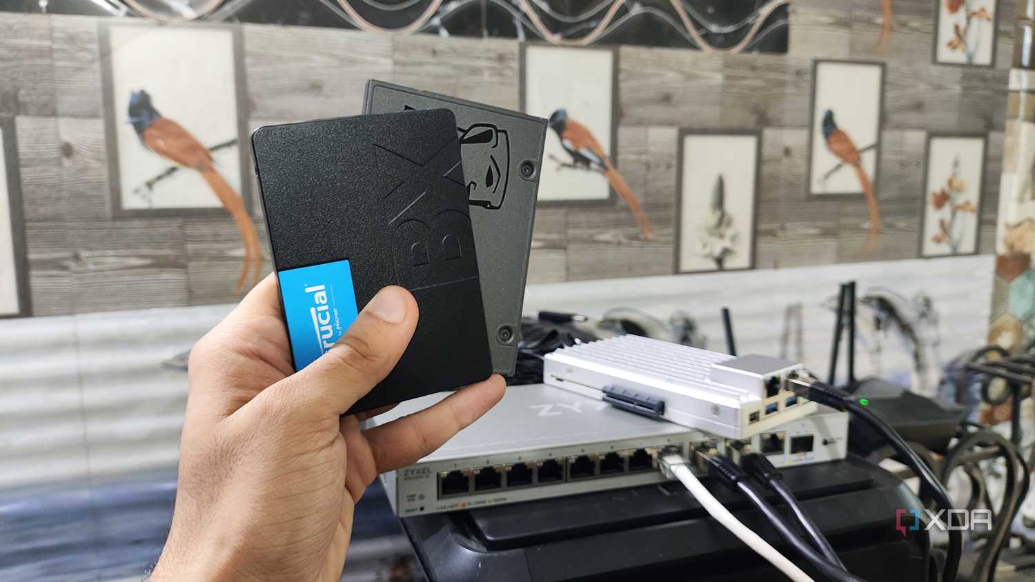A person holding two SSDs, with a ZimaBoard 2 lying in the background